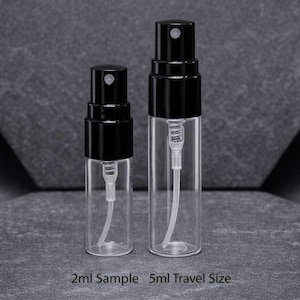 May include: Two clear glass spray bottles with black caps. The bottles are labeled as 2ml Sample and 5ml Travel Size. The bottles have a white internal tube and spray mechanism. The background is a dark gray surface.
