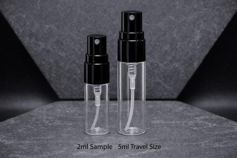 May include: Two clear glass spray bottles with black caps. The bottles are labeled as 2ml Sample and 5ml Travel Size. The bottles have a white internal tube and spray mechanism. The background is a dark gray surface.