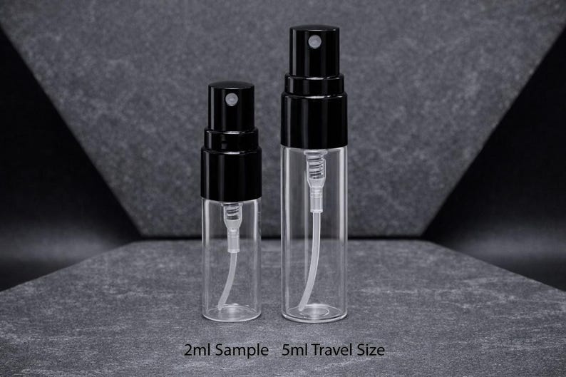 May include: Two clear glass spray bottles with black caps. The bottles are labeled as 2ml sample and 5ml travel size. The bottles have a white internal tube and spray mechanism. The background is a dark gray surface.