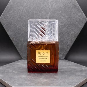May include: A brown perfume bottle with a clear, textured cap. The bottle has a gold label with Arabic text and the words "KHAMRAH DUKHAN Lattafa". The bottle is displayed on a gray surface against a gray background.