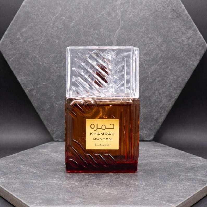 May include: A brown perfume bottle with a clear, textured cap. The bottle has a gold label with Arabic text and the words "KHAMRAH DUKHAN Lattafa". The bottle is displayed on a gray surface against a gray background.