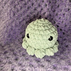 May include: A light green crocheted octopus with black button eyes. The plush toy is made of a soft, textured yarn and sits on a purple and white crocheted blanket. The octopus has eight small tentacles.