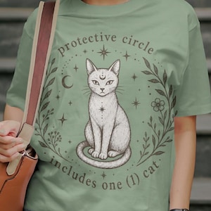 Puede incluir: Camiseta verde salvia con una ilustración detallada de un gato blanco dentro de un círculo decorativo. El diseño incluye estrellas, una luna creciente y elementos botánicos. El texto dice "protective circle includes one (1) cat."