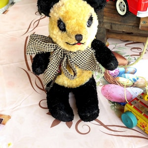 May include: A handmade panda bear plush toy with a yellow face and black ears, arms, and legs. The bear has a checkered bow tie and a small red mouth. The plush is on a patterned surface with other toys in the background.