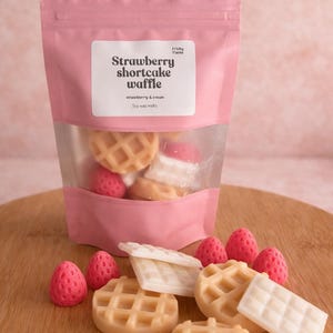 Puede incluir: Una bolsa rosa de ceras de soja con la etiqueta "Strawberry Shortcake Waffle" con aromas de fresa y crema. La bolsa tiene una ventana transparente que muestra ceras con forma de gofre y fresa. Varias ceras están esparcidas sobre una superficie de madera.