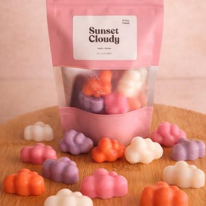 Puede incluir: Una bolsa rosa con una ventana transparente muestra ceras aromáticas en forma de nube de varios colores. La etiqueta dice "Sunset Cloudy" con "Vanilla-Mimosa" y "Soy Wax Melts" debajo. Varias ceras en forma de nube están dispersas.