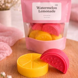 Puede incluir: Una bolsa rosa con el texto "Watermelon Lemonade" contiene ceras aromáticas con forma de rodajas de sandía y limón. Una rodaja de limón amarilla y una rodaja de sandía roja están delante de la bolsa. Las ceras están sobre una superficie de madera.