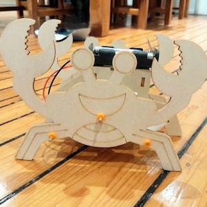 Peut inclure: Un robot en forme de crabe en bois avec un visage souriant, des pinces et des pattes. Le robot a un bloc-piles et un petit moteur attach&eacute;s. Le crabe est en bois de couleur claire et a des accents orange. Il est pos&eacute; sur un sol en bois.