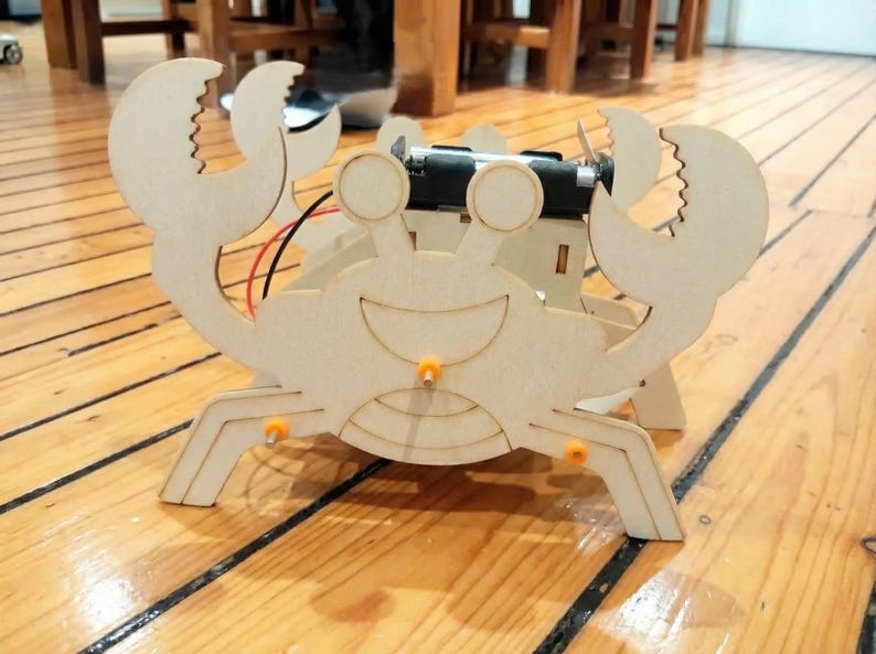 Peut inclure: Un robot en forme de crabe en bois avec un visage souriant, des pinces et des pattes. Le robot a un bloc-piles et un petit moteur attach&eacute;s. Le crabe est en bois de couleur claire et a des accents orange. Il est pos&eacute; sur un sol en bois.
