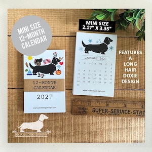 May include: A 2027 mini 12-month calendar featuring a long hair dachshund design. The calendar measures 2.17 inches by 3.35 inches as shown by a wooden ruler. The cover displays a black dog with seasonal icons, and the interior page shows January 2027.