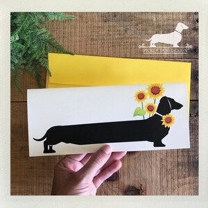 May include: A white card featuring a black dachshund silhouette adorned with sunflowers. The card is held in a hand, with a yellow envelope behind it. The background is a wooden surface, and the card has the text "PICKLE DOG DESIGN".