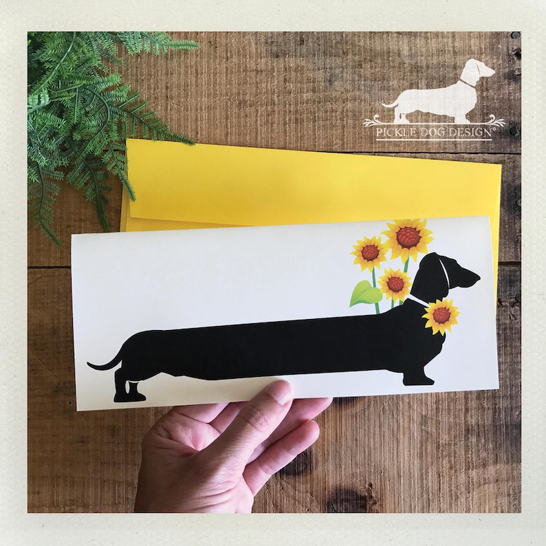 May include: A white card featuring a black dachshund silhouette adorned with sunflowers. The card is held in a hand, with a yellow envelope behind it. The background is a wooden surface, and the card has the text "PICKLE DOG DESIGN".