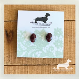 May include: A pair of oval-shaped, dark red earrings on a white card with a dachshund logo and the text "PICKLE DOG DESIGN". The card rests on a wooden surface.