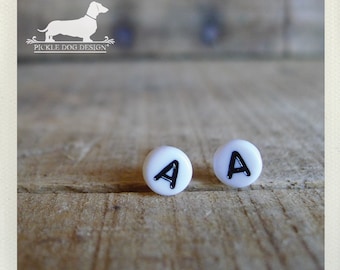 May include: A pair of white stud earrings, each featuring the black letter "A". The earrings are set against a rustic wooden surface. The image also includes a logo with a dachshund silhouette and the text "PICKLE DOG DESIGN".