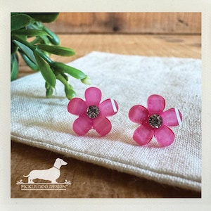May include: A pair of pink flower-shaped stud earrings with a clear rhinestone center and a white stripe on one petal. The earrings are displayed on a beige linen surface, with green foliage in the background.