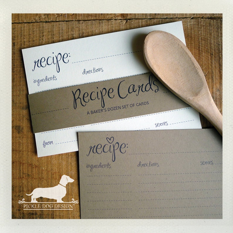 May include: Recipe cards with a wooden spoon on a wooden surface. The cards are white and tan with the word "recipe" in a handwritten font. The cards have lines for ingredients, directions, and servings. The text "Recipe Cards" is also visible.