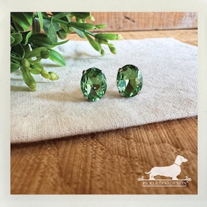 May include: A pair of oval-shaped stud earrings featuring faceted light green stones set in silver-toned metal. The earrings are displayed alongside a white logo graphic of a dachshund with the text PICKLE DOG DESIGN at the bottom right.