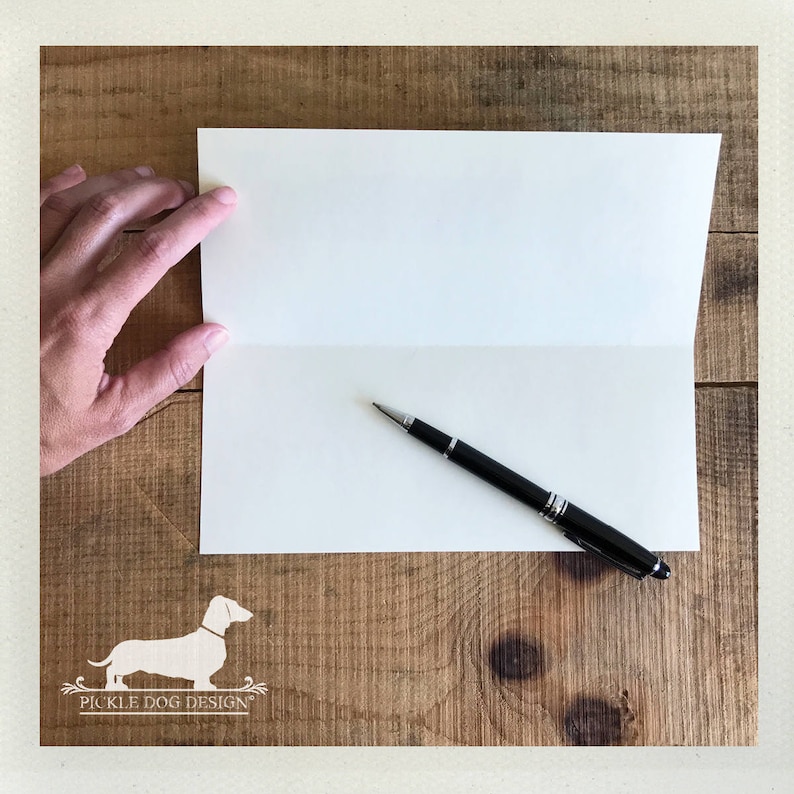 May include: An open, off-white greeting card with a black pen resting on the inside. A hand is holding the card open. The card is on a wooden surface. The Pickle Dog Design logo is in the bottom left corner.