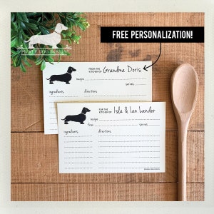 May include: Two recipe cards with a dachshund silhouette, one labeled "From the Kitchen of Grandma Doris" and the other "For the Kitchen of Isla & Ian Lander." The cards have spaces for ingredients, directions, and servings. The image also includes a wooden spoon and the text "FREE PERSONALIZATION!"
