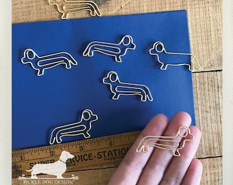 May include: Gold-colored dachshund-shaped paper clips arranged on a blue surface. The paper clips are made of wire and have a simple, stylized design. The image also includes a ruler and the "Pickle Dog Design" logo.