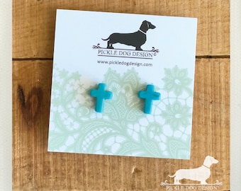 May include: Turquoise cross-shaped stud earrings displayed on a card with a dachshund logo and the text "Pickle Dog Design". The card has a light green floral pattern. The earrings are on a wooden surface.