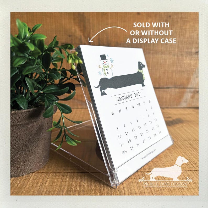 May include: A January 2027 desk calendar featuring a black dachshund wearing a green scarf with a snowman on its back, displayed in a clear plastic case. The text reads SOLD WITH OR WITHOUT A DISPLAY CASE and includes the website address.