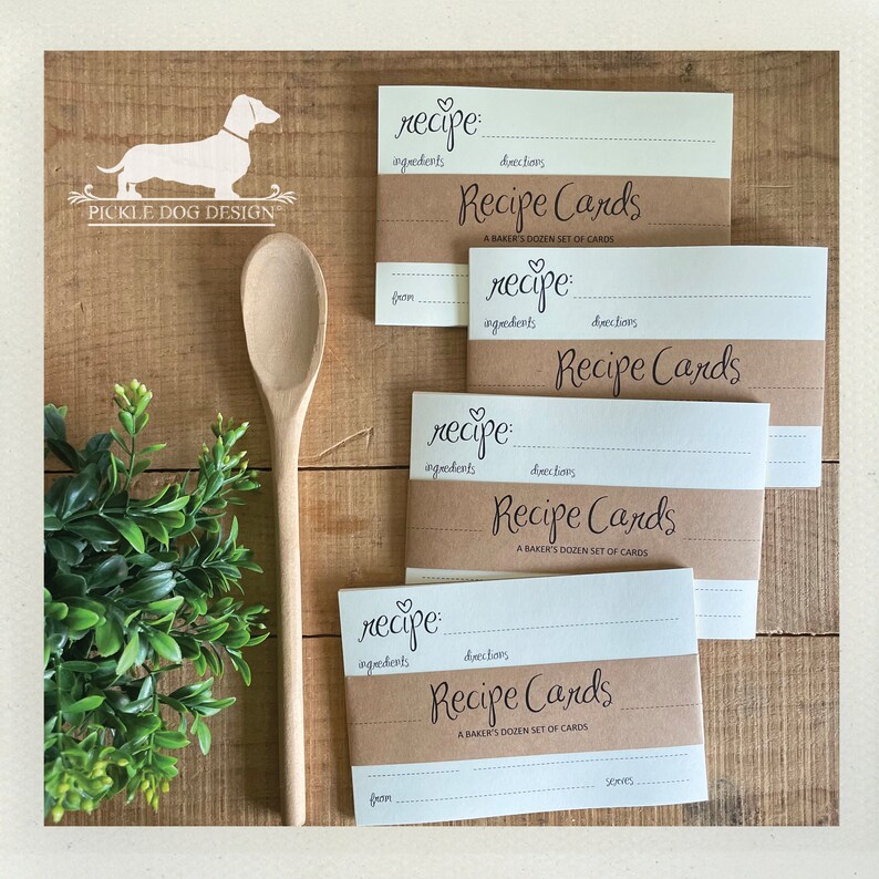 May include: A wooden spoon rests beside a stack of recipe cards. The cards have a cream-colored top and bottom with a brown band in the middle. The text "Recipe Cards" and "A Baker's Dozen Set of Cards" are printed on the cards.