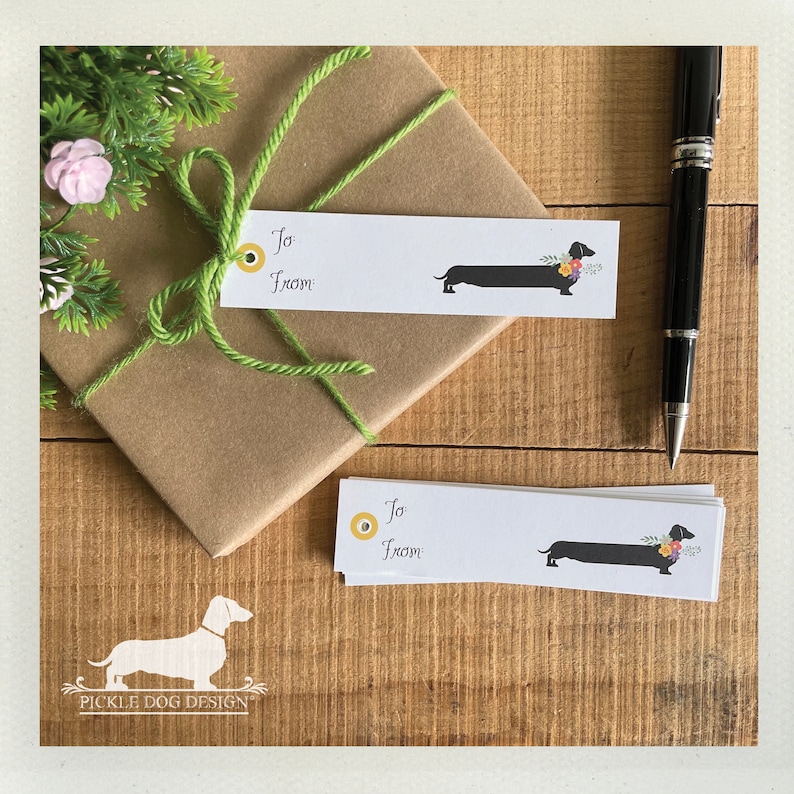 Pu&ograve; includere: Bigliettini regalo bianchi con un'illustrazione di bassotto e le parole "To From". I bigliettini presentano un disegno di bassotto nero con accenti floreali. Un regalo confezionato con spago verde e una penna sono anche nell'immagine.