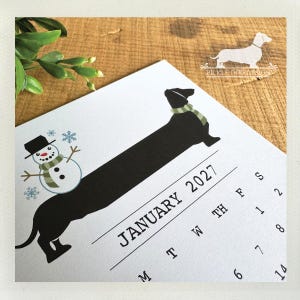 May include: A white calendar page for January 2027 featuring a black dachshund silhouette wearing a green and white striped scarf. A small snowman with a matching scarf and black hat sits on the dog's back. The text reads January 2027.