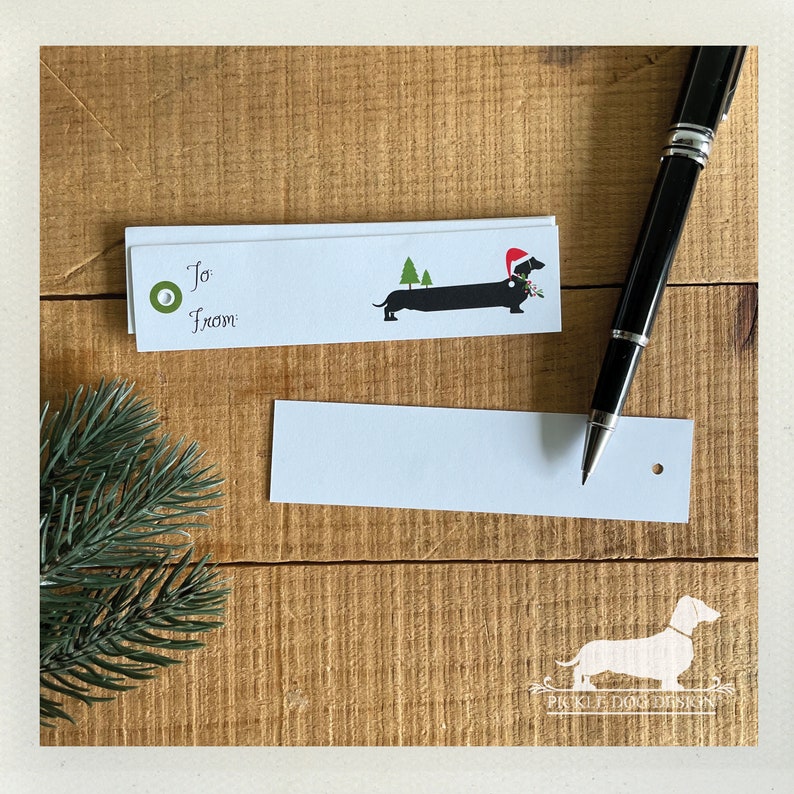 Pu&ograve; includere: Bigliettini regalo bianchi con un'illustrazione di bassotto con cappello da Babbo Natale e rametto di agrifoglio. I bigliettini riportano le parole "A" e "Da". Una penna nera &egrave; appoggiata su una superficie di legno.