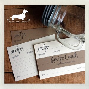 May include: Recipe cards with a heart above the word "recipe" in a handwritten font. The cards have lines for ingredients, directions, and a "from" section. A glass jar with a handle is in the background. The cards are on a wooden surface.