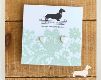 Könnte beinhalten: Weiße herzförmige Ohrstecker auf einer Karte mit einem Dackel-Logo und dem Text "Pickle Dog Design". Die Karte hat ein hellgrünes Spitzendesign. Der Hintergrund ist eine Holzoberfläche.