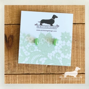 May include: A pair of light green rectangular stud earrings on a white card with a dachshund logo and the text "Pickle Dog Design". The card has a light green lace pattern. The earrings are displayed on a wooden surface.