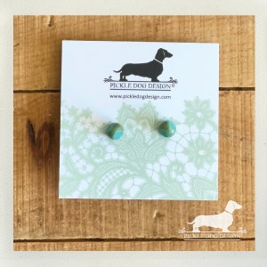 May include: A pair of turquoise and brown stud earrings on a white card with a lace pattern and the text "PICKLE DOG DESIGN". The card is on a wooden surface. The earrings are small, round, and have a marbled appearance.