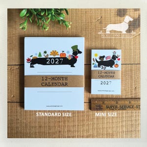 May include: Two 2027 12-month calendars with a dachshund illustration. The larger calendar features a dachshund with seasonal icons and the year 2027. The smaller calendar shows a dachshund with a flag and icons. A ruler shows a 3-inch width.