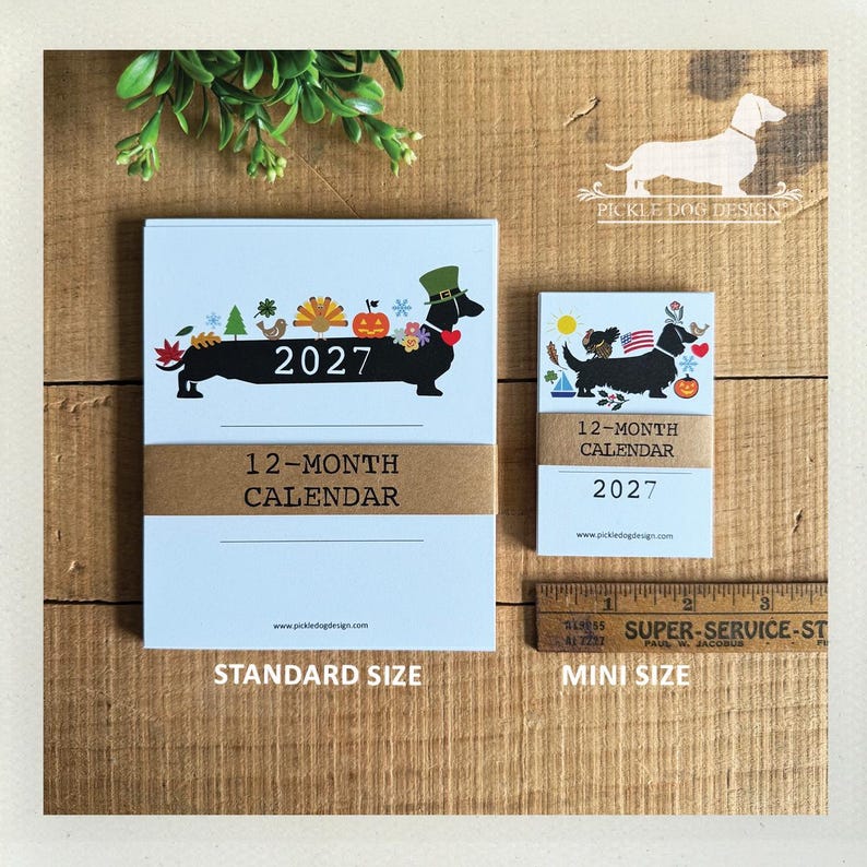 May include: Two 2027 12-month calendars with a dachshund illustration. The larger calendar features a dachshund with seasonal icons and the year 2027. The smaller calendar shows a dachshund with a flag and icons. A ruler shows a 3-inch width.