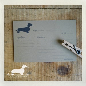May include: A recipe card with a dachshund silhouette, recipe, ingredients, directions, and serves sections. A wooden clothespin rests on the card. The card is on a wooden surface.