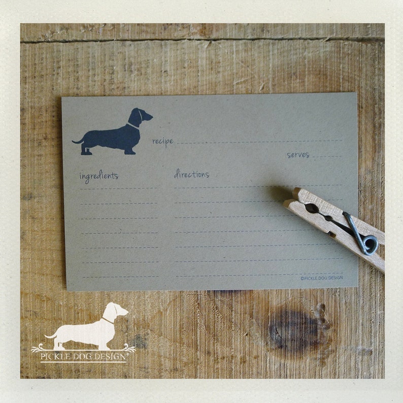 May include: A recipe card with a dachshund silhouette, recipe, ingredients, directions, and serves sections. A wooden clothespin rests on the card. The card is on a wooden surface.