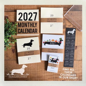 May include: A collection of 2027 monthly calendars featuring dachshund illustrations. The set includes various paper calendars with brown paper bands, a long vertical black bookmark calendar, and a small card, all displaying the year 2027.