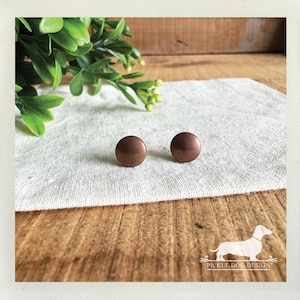 May include: Two round, copper-colored stud earrings sit on a white linen cloth. The earrings are smooth and have a slight dome shape. The background includes a wooden surface, green foliage, and a logo.