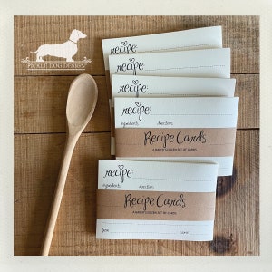 May include: A stack of recipe cards with a brown band that reads "Recipe Cards: A Baker's Dozen Set of Cards." The cards have space for ingredients and directions. A wooden spoon and a logo of a dachshund are also in the image.