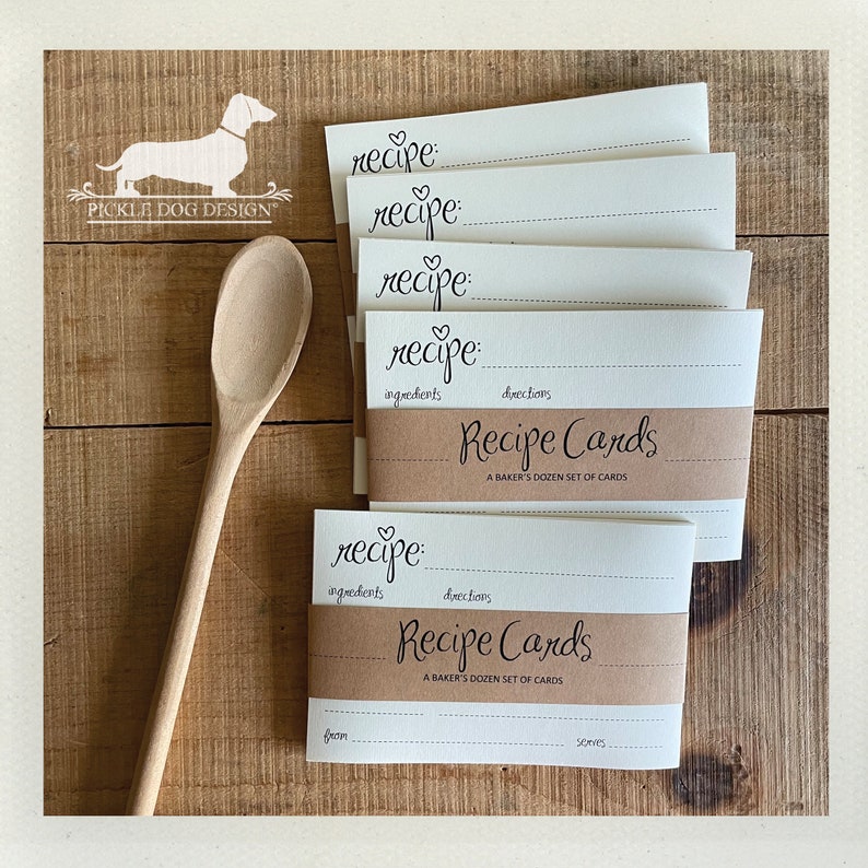 May include: A stack of recipe cards with a brown band that reads "Recipe Cards: A Baker's Dozen Set of Cards." The cards have space for ingredients and directions. A wooden spoon and a logo of a dachshund are also in the image.