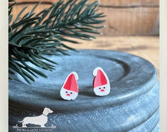 May include: Two Santa Claus-shaped stud earrings with red hats and white faces. The earrings are on a gray, textured surface with a sprig of evergreen and the Pickle Dog Design logo in the background.