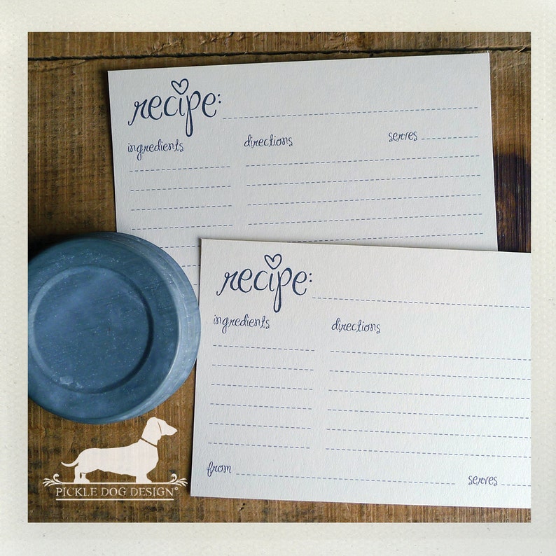 May include: Two white recipe cards with blue handwritten text and a blue heart above the word "recipe". The cards have lines for ingredients, directions, and servings. A blue plate and a dog logo are also visible.