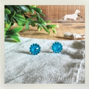 Op de afbeelding: Twee turquoise oorstekers met een gestructureerd, kralenontwerp. De oorbellen zijn rond en hebben een sprankelend midden. Ze worden gepresenteerd op een lichtgrijze linnen doek, met groen en een houten oppervlak op de achtergrond.