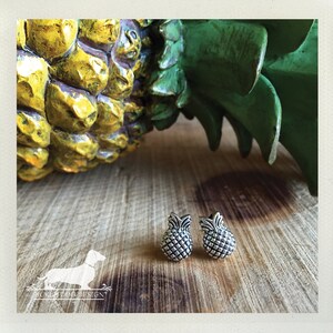 May include: A pair of silver-colored pineapple stud earrings. The earrings are detailed with a textured surface, mimicking the skin of a pineapple. The earrings are displayed on a wooden surface, with a yellow and green pineapple in the background.
