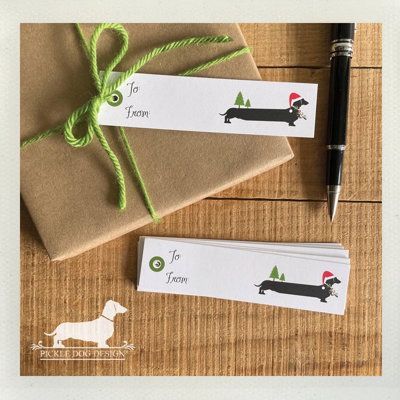 Pu&ograve; includere: Bigliettini regalo bianchi con un bassotto con cappello di Babbo Natale e rametto di agrifoglio. Le etichette hanno le parole "To" e "From". Un regalo avvolto con spago verde e una penna nera sono anche nell'immagine.