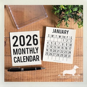 May include: A 2026 monthly calendar and a January calendar card sit on a wooden surface. The 2026 calendar card has the text "2026 MONTHLY CALENDAR" in bold black font. A black pen and a clear plastic container are also present.