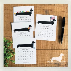 May include: Four white calendar pages for April, May, June, and July 2027, each featuring a black dachshund illustration. The dogs are decorated with colorful birds, flowers, a blue striped tie, or an American flag. A black pen lies to the right.