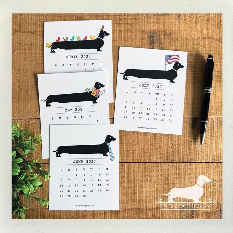 May include: Four white calendar pages for April, May, June, and July 2027, each featuring a black dachshund illustration. The dogs are decorated with colorful birds, flowers, a blue striped tie, or an American flag. A black pen lies to the right.
